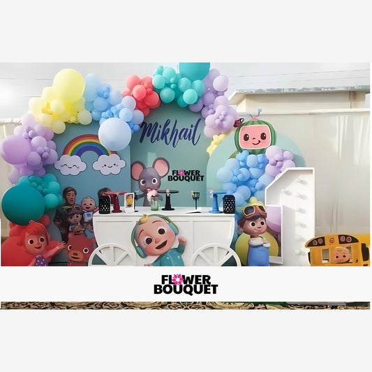 Colorful birthday party decoration with a pastel-colored balloon arch, large 'Celebrant' sign, cute cartoon characters, rainbow with clouds, and a white cart-style table with party items, creating a joyful and festive atmosphere.