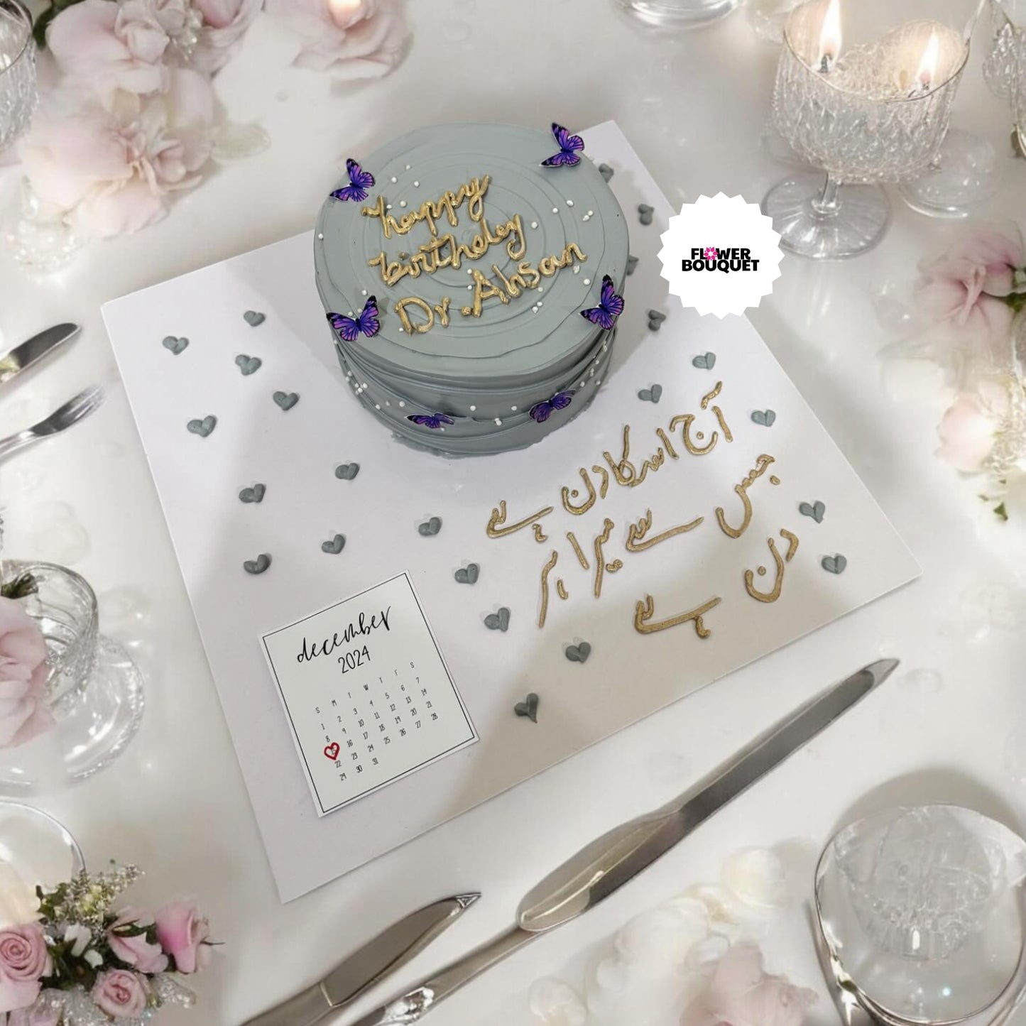 Gray birthday cake with purple butterflies, surrounded by pink flowers, candles, and a calendar on an elegant table setting.