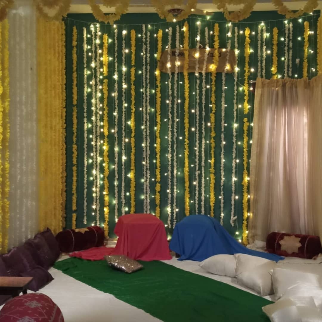 Vibrant yellow marigold floral string decorations with fairy lights hanging on a green fabric backdrop, creating a festive atmosphere for an event.