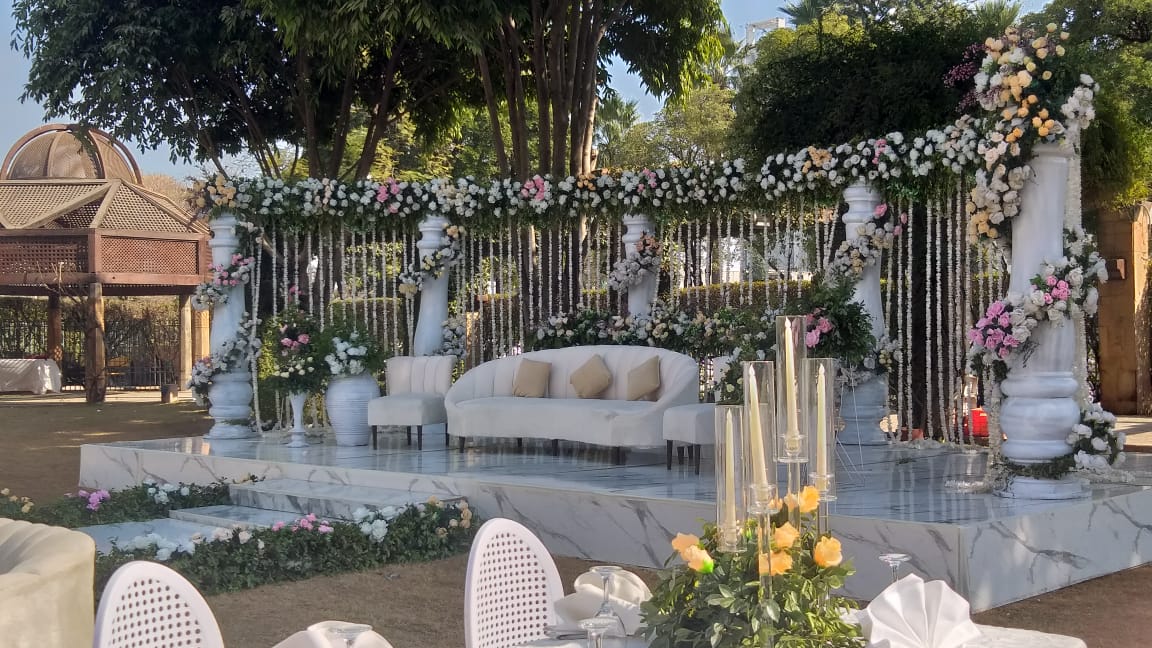 A stunning Walima decoration setup featuring a white and gold theme. The stage includes an ornate upholstered sofa, a floral backdrop with pastel roses and lilies, elegant draped curtains, and soft fairy lights. 