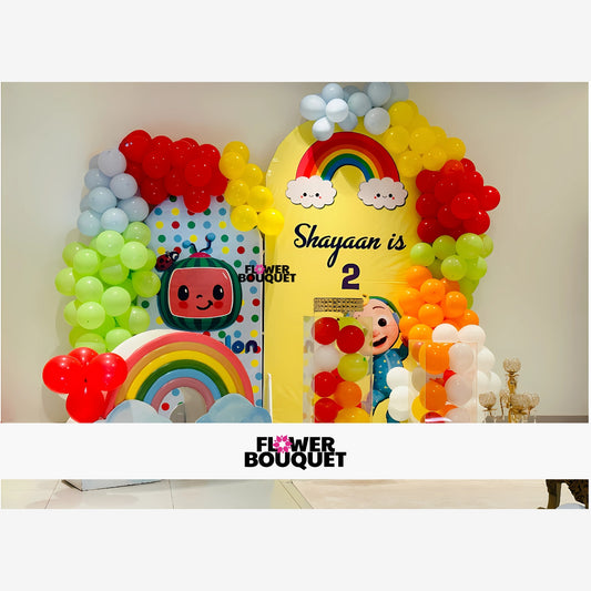 Colorful birthday party decoration with a large yellow arch, rainbow and clouds, 'Happy 2nd Birthday' text, clusters of red, yellow, green, blue, and white balloons, cartoon character, and playful setup creating a vibrant and festive atmosphere.