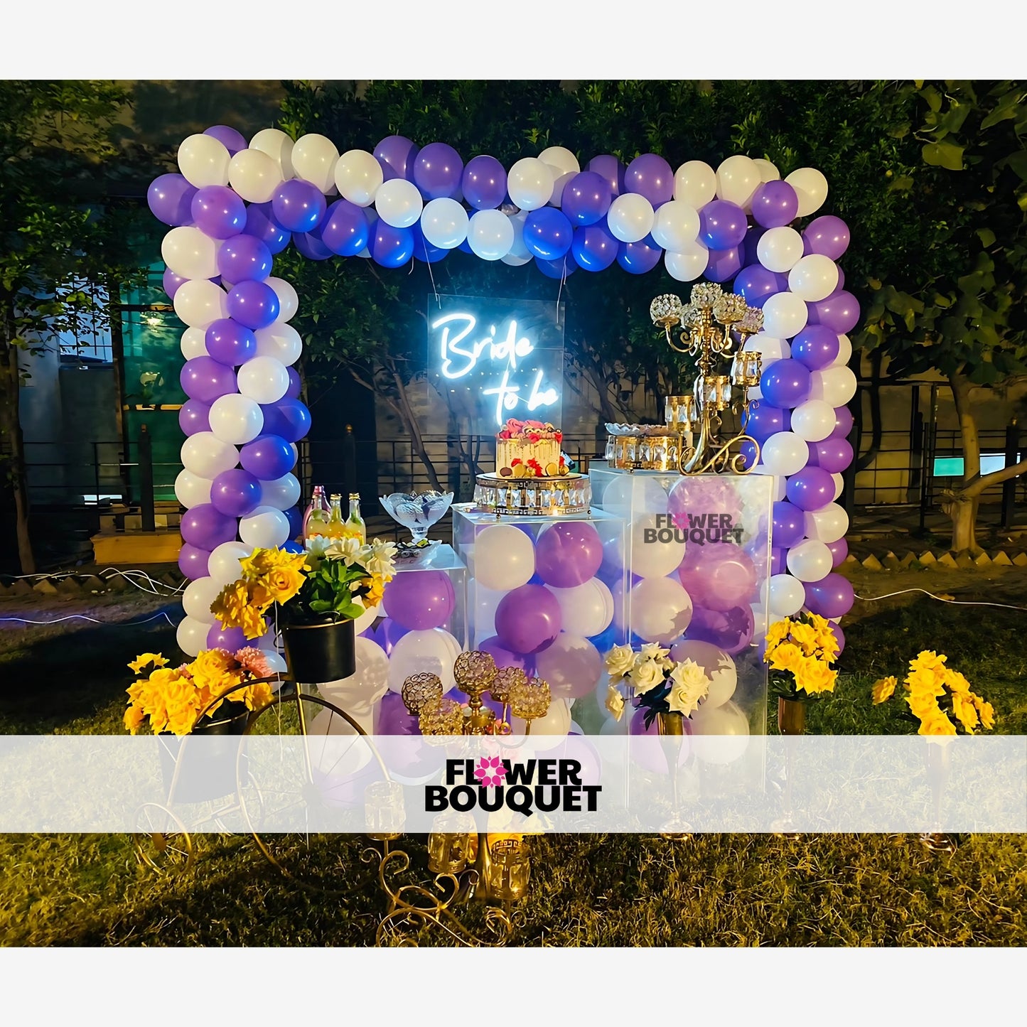 Decorative setup for a bridal event with a balloon arch, neon "Bride to be" sign, and a table with a cake and decorations.