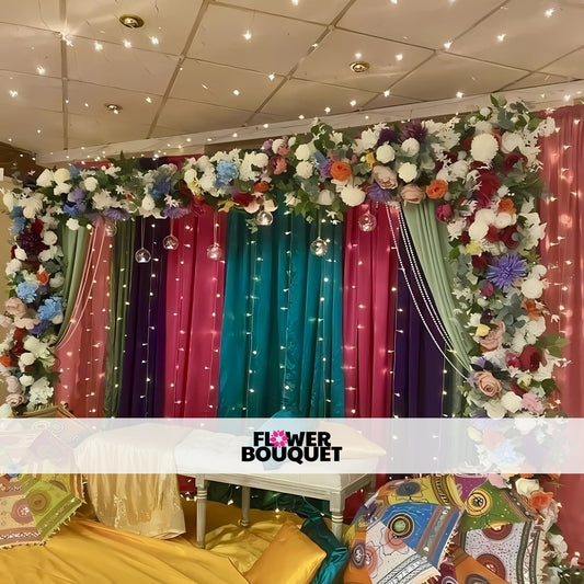 Mehndi backdrop with multicolored drapes, fairy lights, floral arrangements, and decorative umbrellas for an elegant and festive setup.
