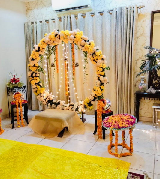 A beautifully decorated circular floral backdrop with yellow and white flowers, string lights, and floral stools, ideal for wedding or mehendi ceremony decor.