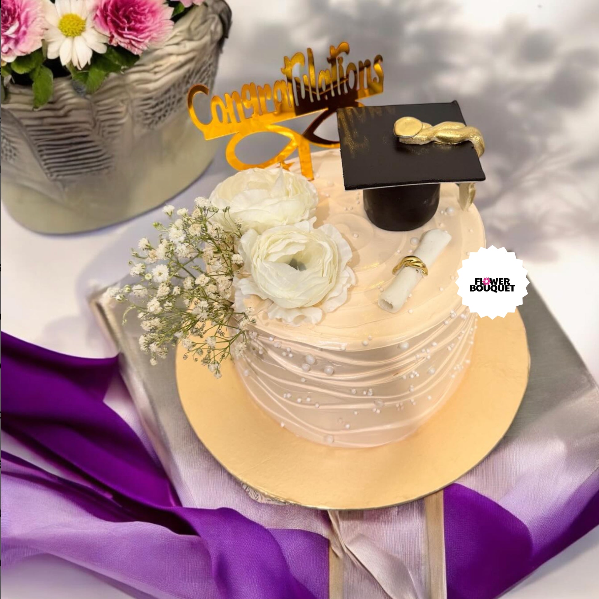 Graduation cake with mini cap, diploma, and flowers, next to a flower basket. Gold "Congratulations" sign on top.