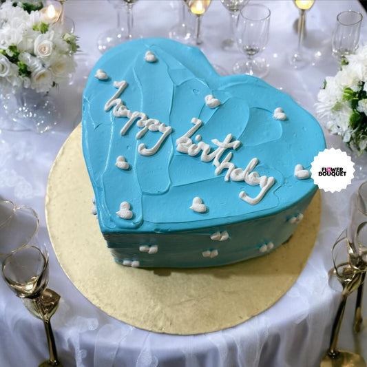 Heart-shaped blue birthday cake with white lettering, on gold board, surrounded by glasses and flowers.