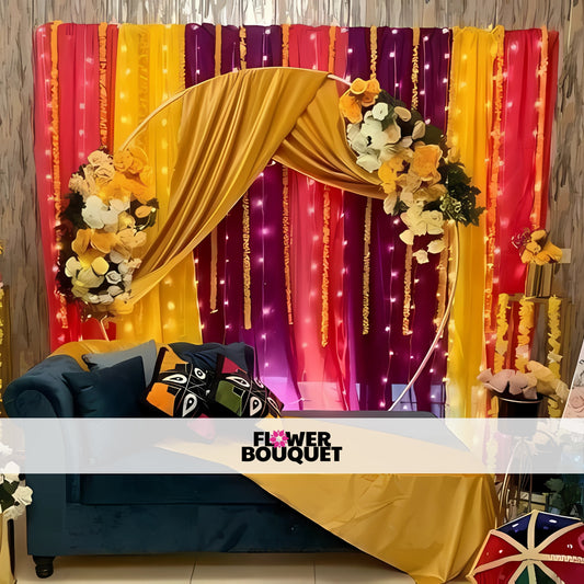 Mehndi decor with a circular floral frame, yellow and white flowers, colorful drapes, fairy lights, and a modern blue sofa with vibrant cushions.