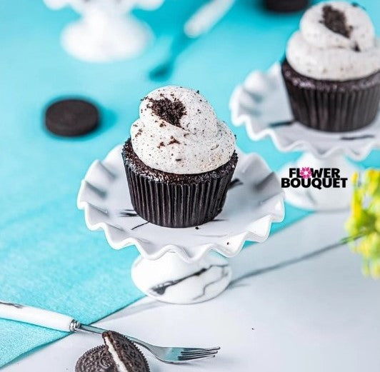 A beautifully presented Oreo Bliss Cupcake on a marble-patterned pedestal stand, topped with creamy Oreo frosting and crushed Oreo crumbs, with a bright blue background and decorative table setting.