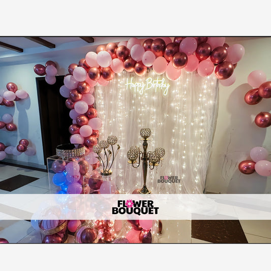 Birthday party setup with a pink and rose gold balloon arch, white sheer curtains, and string lights.