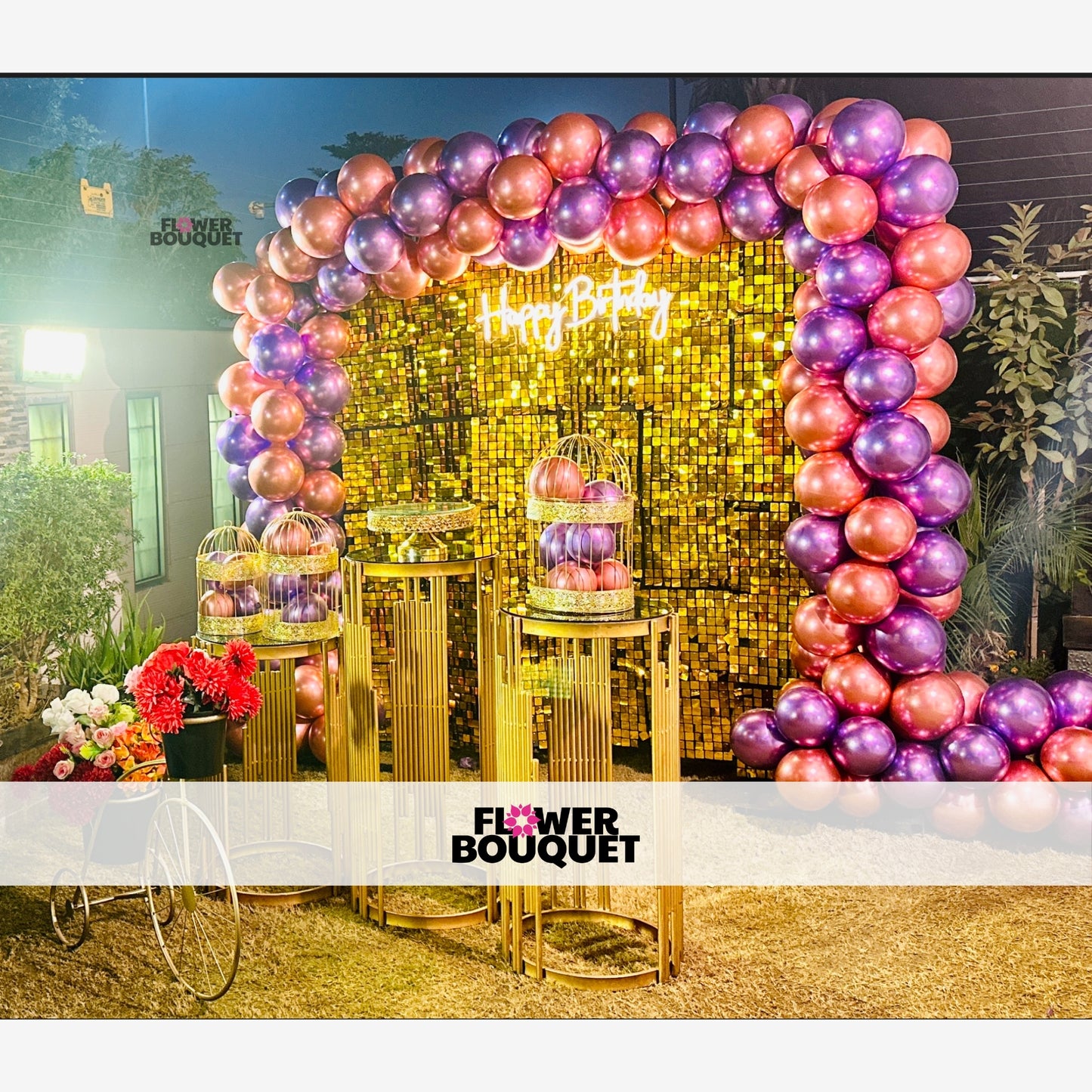 Decorative setup for a birthday celebration with a large balloon arch, golden sequin backdrop, and floral arrangements.