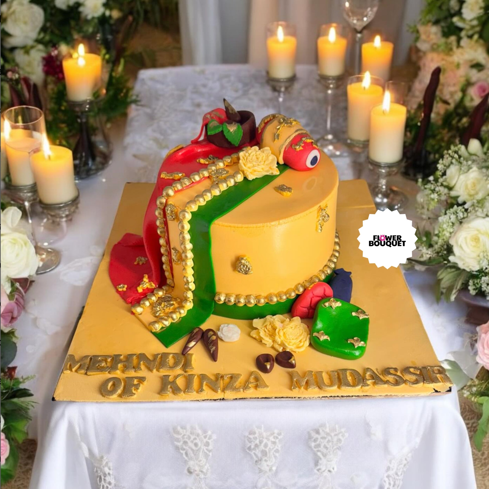 Colorful cake with fruit and jewelry decorations, gold beads, and "MEHNDI OF KINZA MUDASSIR" text, surrounded by candles and flowers.