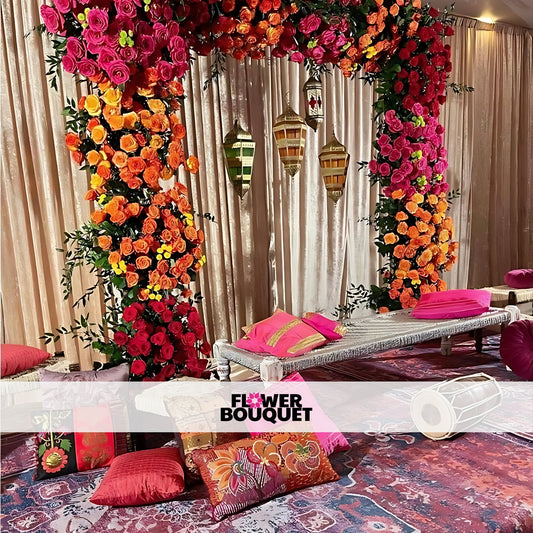 A colorful Mehndi stage setup with pink, orange, and yellow floral arrangements, metallic lanterns, traditional low seating, and vibrant cushions on an ornate carpet.